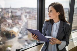 An investor holds a clipboard while walking through an office and looking out a window at a city neighborhood.